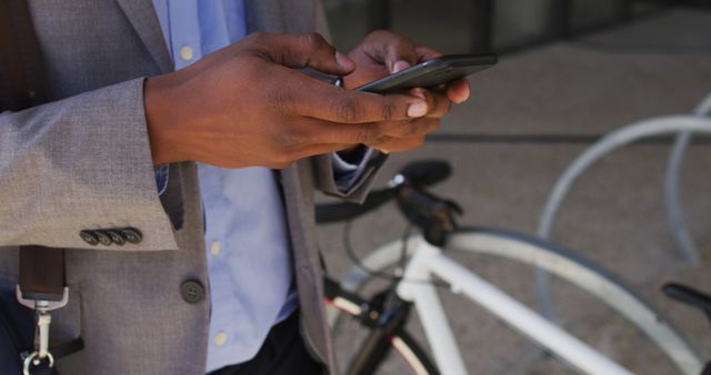 Businessman is standing outdoors, wearing a suit, and checking smartphone next to a bicycle. This can be used for profiles, articles, and advertisements focused on urban professionals, modern commuting solutions, business routines, and technology in professional environments.