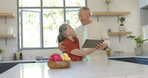 Diverse senior couple embracing and holding tablet in bright modern kitchen