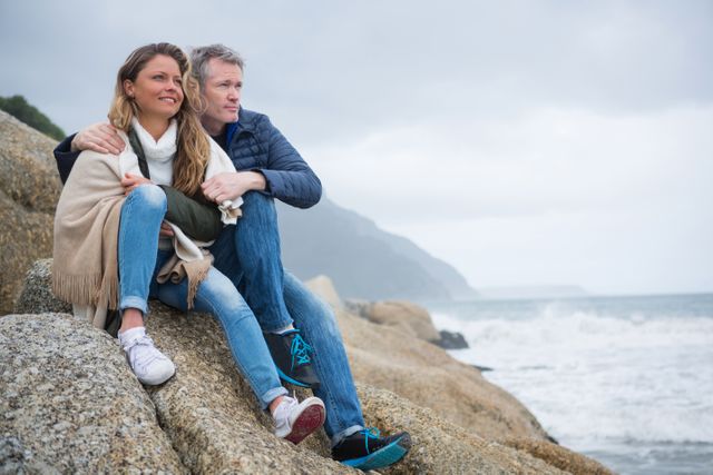 Couple sitting on rocks by the beach, enjoying the scenic ocean view. Ideal for use in travel brochures, romantic getaway promotions, relationship blogs, and lifestyle magazines.