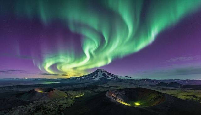 Aurora swirling over volcanic craters and snow-capped stratovolcano under starry sky