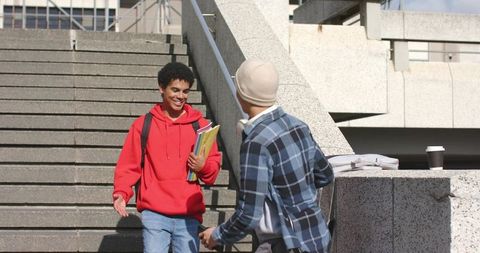 Smiling diverse college friends greeting on campus stairs, red hoodie holding notebooks