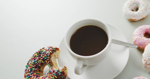 White coffee cup filled with dark coffee placed on white saucer. Partially bitten donut with colorful sprinkles next to cup. Various other sprinkled donuts placed around. Ideal for advertising coffee shops, breakfast menus, dessert parlors, snack posters, brunch gatherings, social media posts.