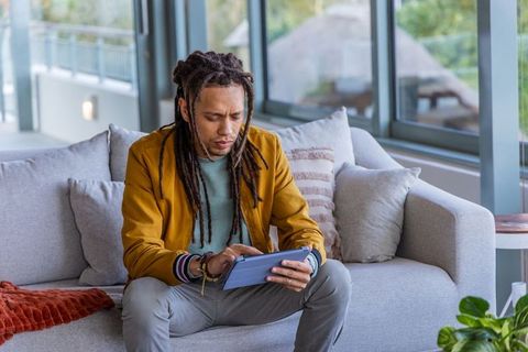 African american man using tablet on gray sofa wearing mustard jacket in modern lounge