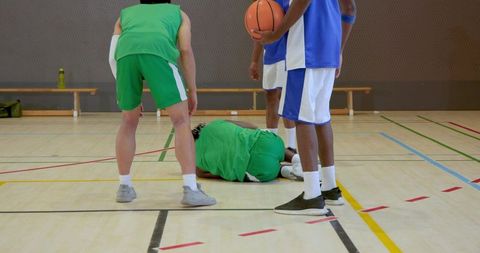 Injured basketball player lying on gym floor with teammates and opponents standing nearby