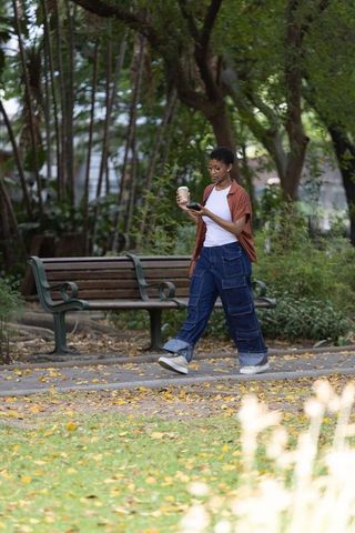 African american woman walking in park holding coffee and phone autumn urban lifestyle