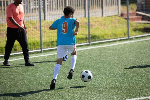 African American boy soccer team player practicing on a green football pitch on a sunny day, running with a ball his male coach in the background. Childhood healthy lifestyle competition.