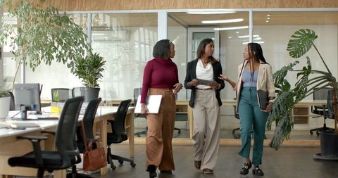 Diverse female coworkers walking and discussing in modern open-plan workspace with plants