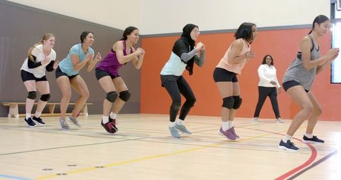 Multicultural women performing squat jumps in indoor gym fitness class with hijab and coach leading
