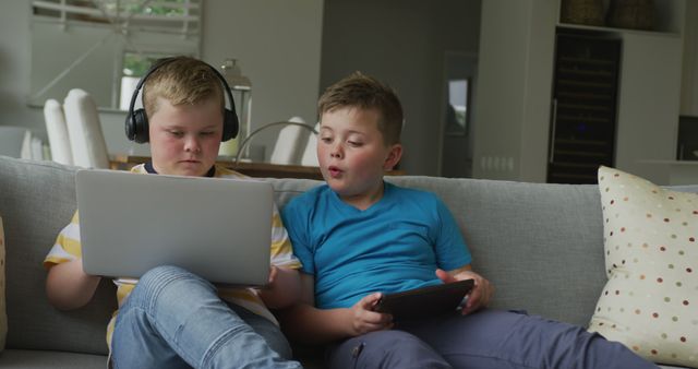 Two boys sitting on a comfortable sofa in a modern living room. One boy is using a laptop and wearing headphones while the other boy is holding a digital tablet and engaging in a conversation. Perfect for themes related to children and technology, learning, family life, and modern lifestyles.