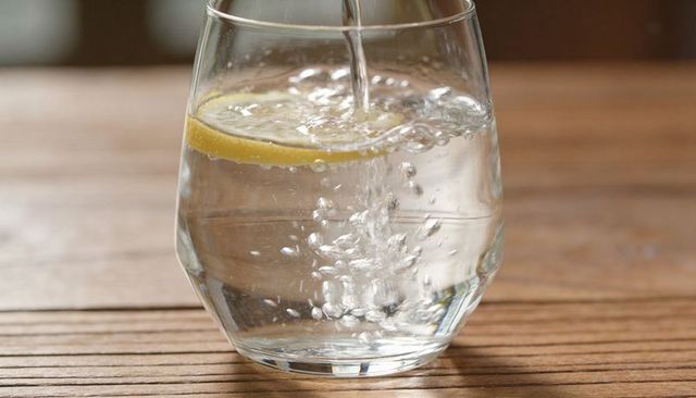 Pouring sparkling water with lemon slice in clear rounded glass on wood table, refreshing