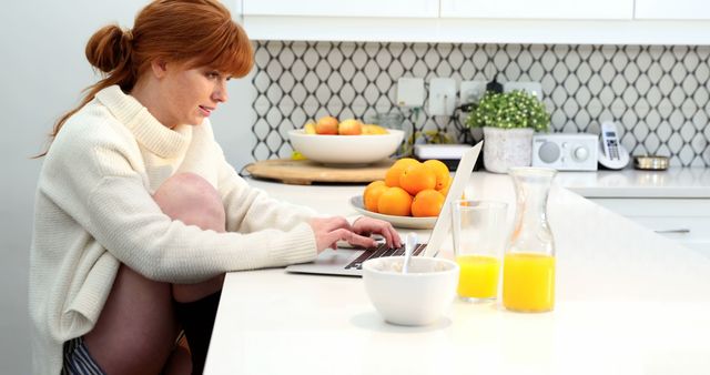 Young woman concentrating on her laptop in a stylish white kitchen with fresh oranges and glasses of orange juice. Ideal for themes like modern work-life balance, remote working, healthy lifestyle, productive mornings, and kitchen design inspiration.