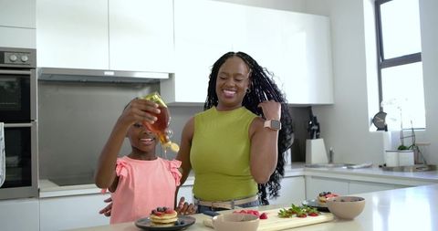 Smiling mother and daughter pouring syrup on pancakes in sunlit modern kitchen