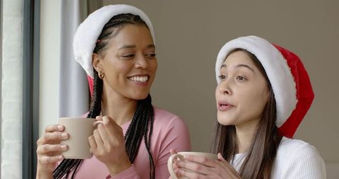 Multiracial friends wearing santa hats enjoying hot drinks by window cozy festive moment