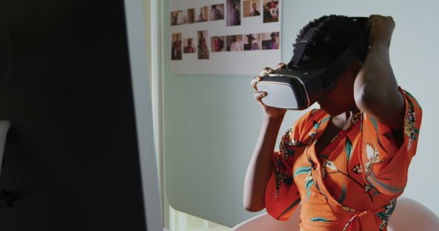 Woman of African descent wearing an orange top, engaging with virtual reality technology by adjusting a VR headset. Ideal for topics on modern technology, innovation, immersive experiences, and digital advancements. Useful for illustrating VR gaming, futuristic studies, and indoor technological environments.