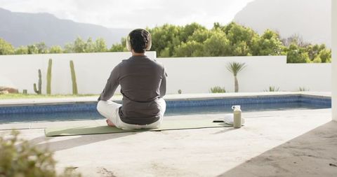 Adult Indian man meditating poolside wearing headphones using laptop for remote work and wellness