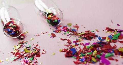 Tipping stemless glasses scattering metallic confetti on pastel pink surface