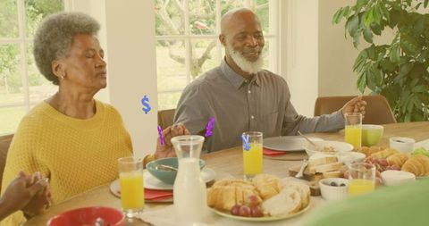 Senior couple holding hands praying at sunlit family breakfast table with croissants and juice