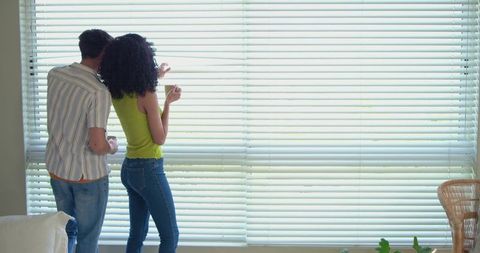 Multicultural couple peeking through horizontal blinds in bright living room sunlight