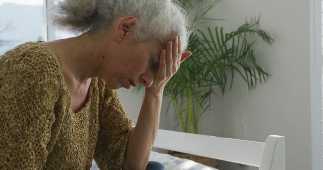 Elderly woman sitting on bed in home bedroom with head in hands, expressing sadness and frustration. Gray-haired woman wears casual brown sweater, natural light from window nearby. Suitable for concepts of mental health, elderly care, emotional stress in aging, and healthcare awareness.