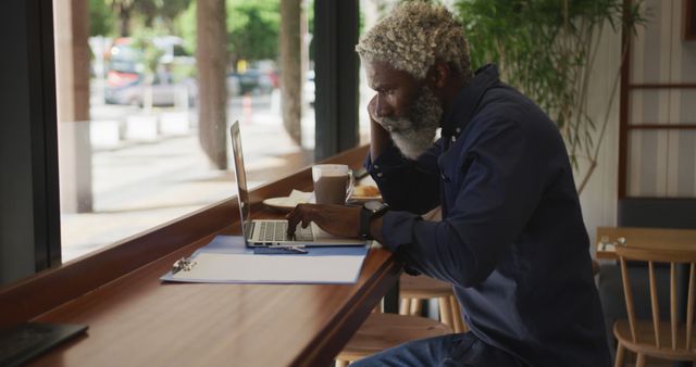 Senior Black man in casual blue jacket working on laptop in a cozy café, concentrating on screen. Takeaway coffee and clipboard on wooden countertop. Ideal for depicting remote work, business productivity, modern technology use, senior professionals, café culture, urban freelancers, and lifestyle.