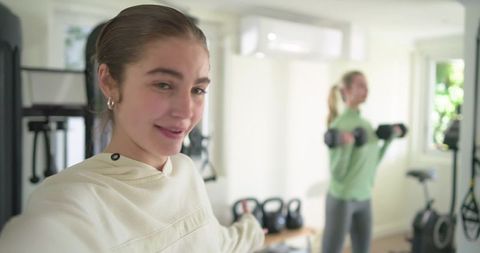Young women taking hoodie selfie while partner lifting dumbbells in bright home gym