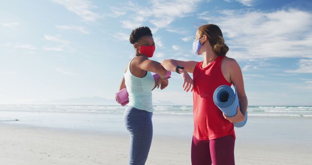 Two friends are greeting each other with elbow bumps on a beach while wearing masks. They are holding yoga mats, suggesting they are about to participate in an outdoor yoga session. The scene promotes a message of social distancing and safe interactions during the pandemic. Useful for content related to health and fitness, pandemic activities, social distancing practices, and outdoor exercise.