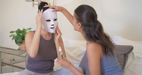 Friends wearing led facial mask and applying serum during cozy at-home skincare routine