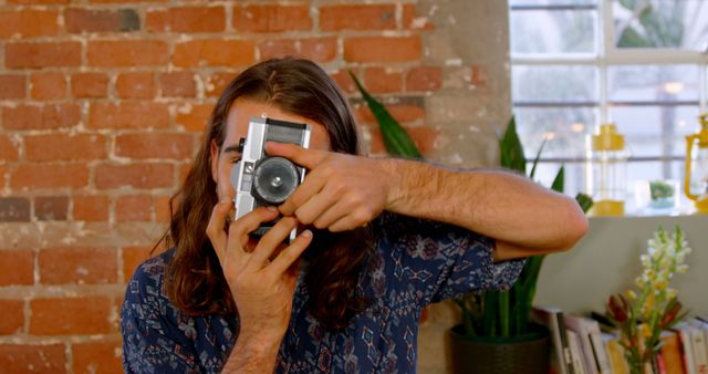 Young man holding a vintage camera while standing in a loft apartment studio with exposed brick walls, greenery in the background, and casual attire. Ideal for themes around photography, hobby, lifestyle, creative photographers, and vintage equipment in modern settings.