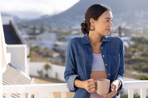 Young woman standing on balcony holding mug, overlooking mountain cityscape at sunrise