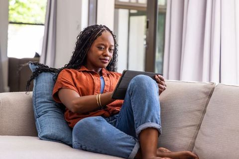 African woman relaxing on couch using tablet at home for reading, streaming, remote work