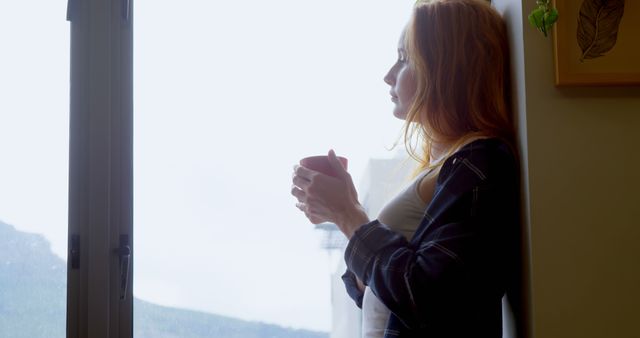 Young Caucasian woman enjoys a quiet moment by the window at home. She sips her coffee, reflecting in the warm morning light.