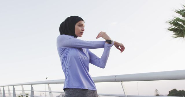 A Muslim woman stretches in an outdoor setting, with athletic clothing and a hijab. She wears a smartwatch, indicating she may be tracking her fitness progress. Suitable for themes on fitness, healthy lifestyle, cultural diversity in sports, and morning routine activities.