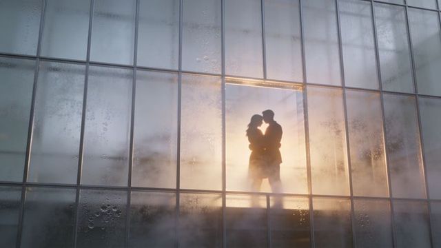 Backlit couple embracing and kissing inside misty glass conservatory silhouette at sunrise