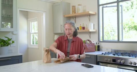 Senior asian woman packing lunch at bright modern kitchen island with smartphone nearby