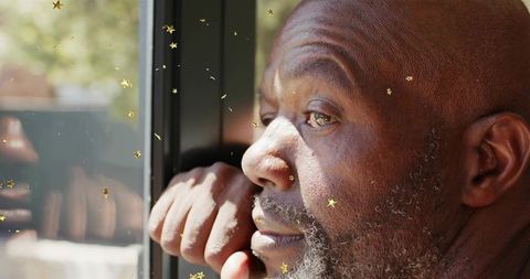 Sunlit senior man gazing through window, resting chin on hand with gold star confetti