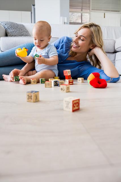 Mother watching her baby boy playing with toys in a cozy living room. The baby is engaged with colorful blocks and a rubber duck, while the mother looks on with a smile. This image is perfect for use in parenting blogs, family lifestyle articles, advertisements for baby products, and educational materials on child development.