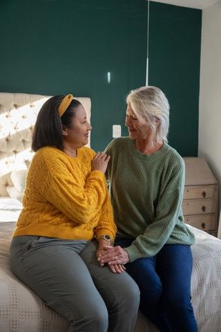Female partners holding hands on bed wearing knit sweaters, intimate cozy home moment