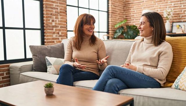 Two friends chatting on cozy sofa wearing turtlenecks and jeans in sunlit living room