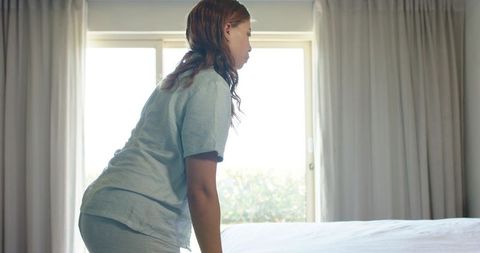 Woman in blue pajamas making bed by backlit sliding door morning minimal bedroom
