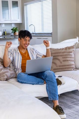 Asian young man celebrating while working on laptop at home on cozy light sectional sofa