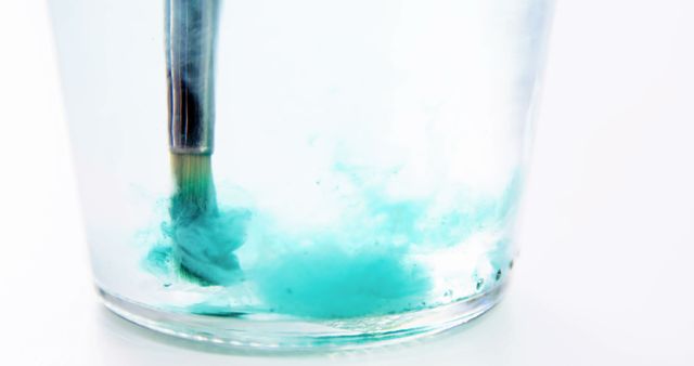 Close-up of a paintbrush dipped in a glass of water with turquoise paint spreading and swirling, ideal for content related to art, creativity, watercolor tutorials, and visual arts concepts. Perfect for illustrating artistic techniques in educational material or promoting art supplies and classes.
