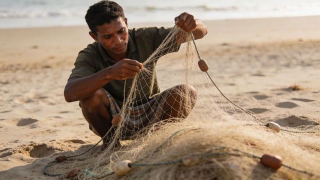 Coastal fisherman repairing traditional net on sunlit sandy beach preparing for fishing