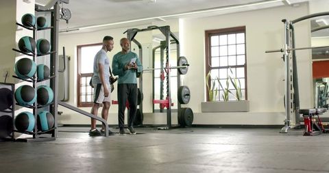 African american trainer coaching client near power rack holding tablet in modern gym