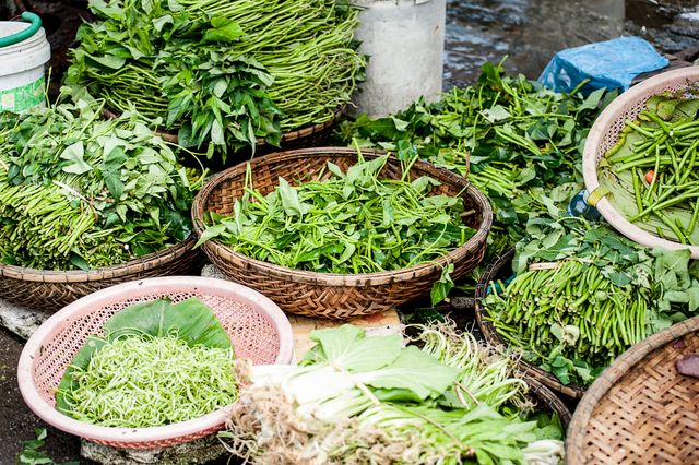 Various types of fresh greens displayed in wicker baskets at outdoor market stall. Ideal for use in articles, blogs, and advertisements about organic produce, local markets, healthy eating, and sustainable living.