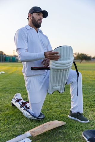 Male cricketer kneeling with batting pads and bat during dusk practice on grassy field