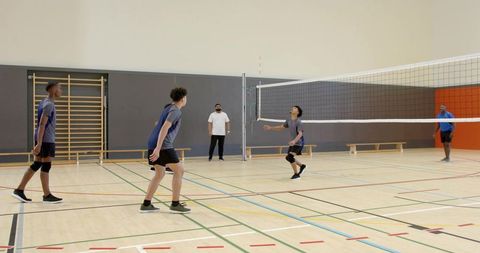 Diverse male volleyball team practicing drills in indoor gym with coach supervising