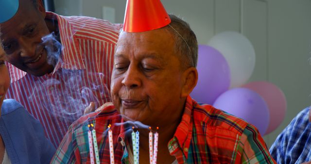 A senior man is blowing out candles on a birthday cake, surrounded by diverse friends or family celebrating with him. Capturing a moment of joy and celebration, the image reflects a warm, festive atmosphere during a special occasion.