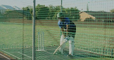 Cricketer practicing in outdoor net wearing helmet, pads and gloves on sunny field