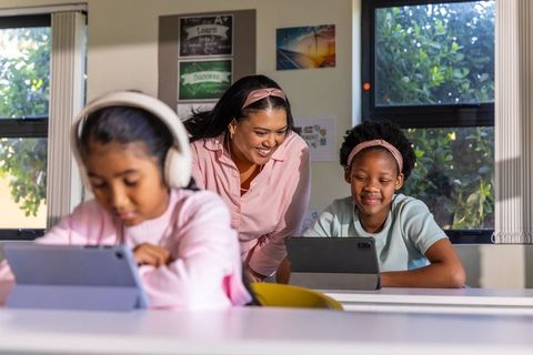 Diverse teacher mentoring students using tablets and headphones in sunlit classroom