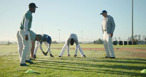 Male Baseball Players Stretching at Dawn while Coach Overseeing Field Practice and Warmups